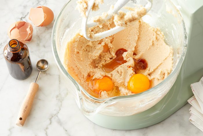 In a large bowl, beat butter and sugars until blended. Beat in eggs and vanilla. Overhead step-by-step image of softened butter, brown sugar and granulated sugar creamed together in a glass mixing bowl. Eggs and vanilla extract are added to the mixture on a light marble surface, with baking tools nearby. This image highlights the beginning stage of making Easter Chocolate Chip Cookies.