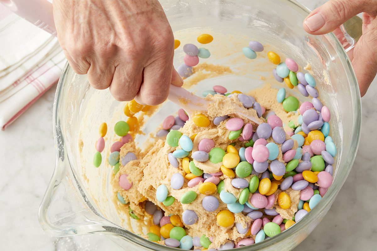 Overhead step-by-step image of pastel milk chocolate M&M’s being folded into thick cookie dough inside a glass bowl. Hands gently mix the colorful candies evenly throughout the batter, creating a festive Easter appearance.
