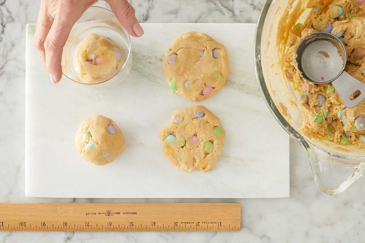Shape 1/4 cupfuls of dough into balls. Flatten each to 3/4-in. thickness (2-1/2-in. diameter), smoothing edge as necessary. Overhead step-by-step image of rounded scoops of Easter cookie dough placed evenly on parchment-lined baking sheets. The pastel M&M’s are visible on top of each dough ball before baking.