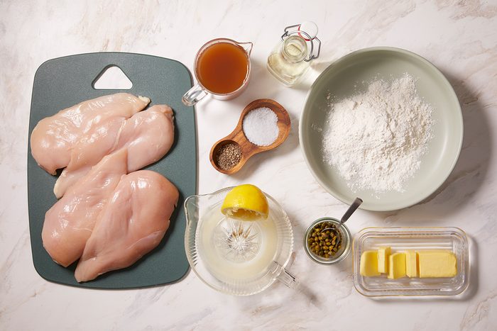 Overhead view of raw chicken breasts on a cutting board, with bowls of flour, butter, capers, lemon juice, chicken broth, oil, salt, and pepper arranged on a white surface.