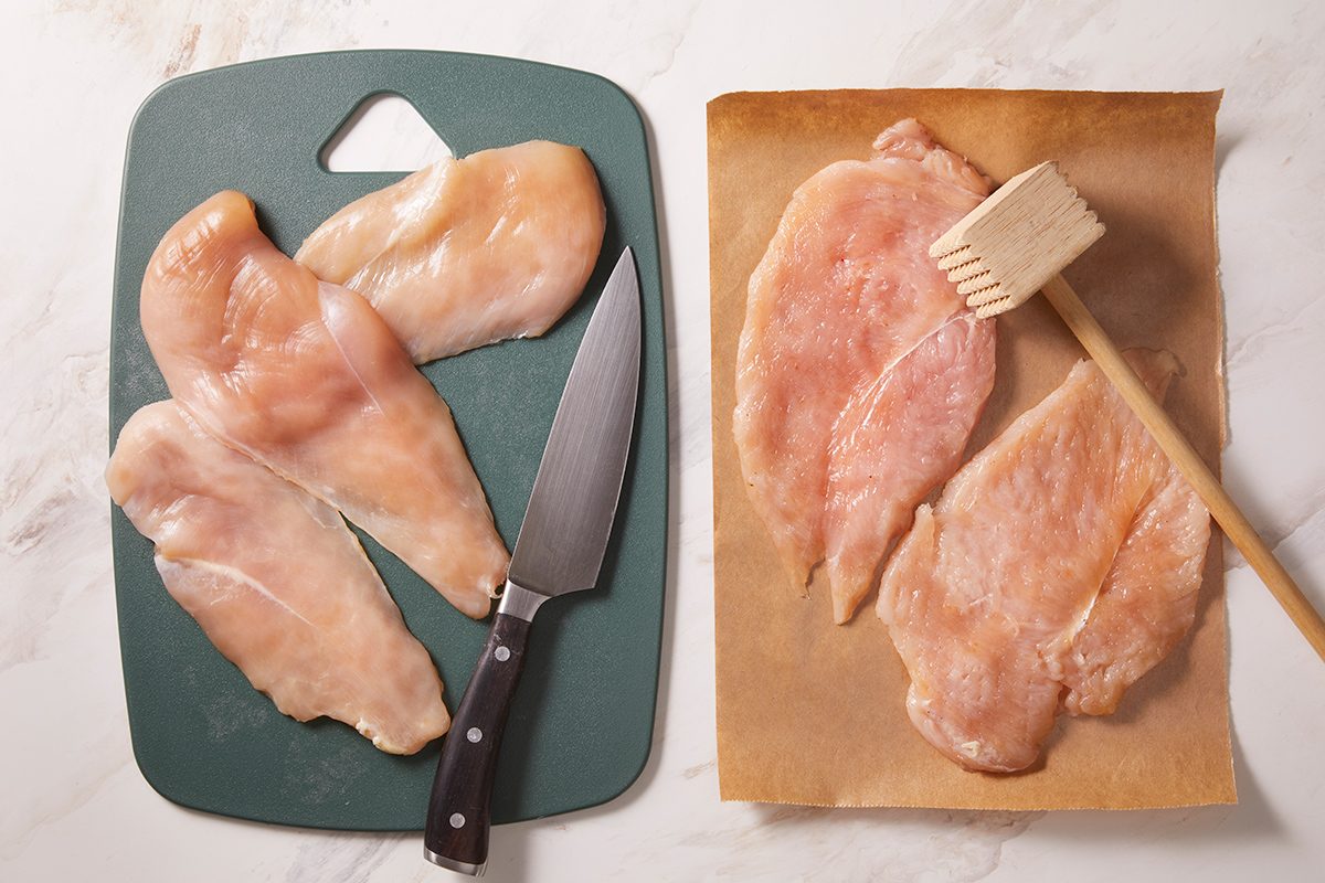 Four raw chicken breasts on a green cutting board with a knife, and two flattened chicken breasts on parchment paper beside a meat tenderizer, all on a white countertop.