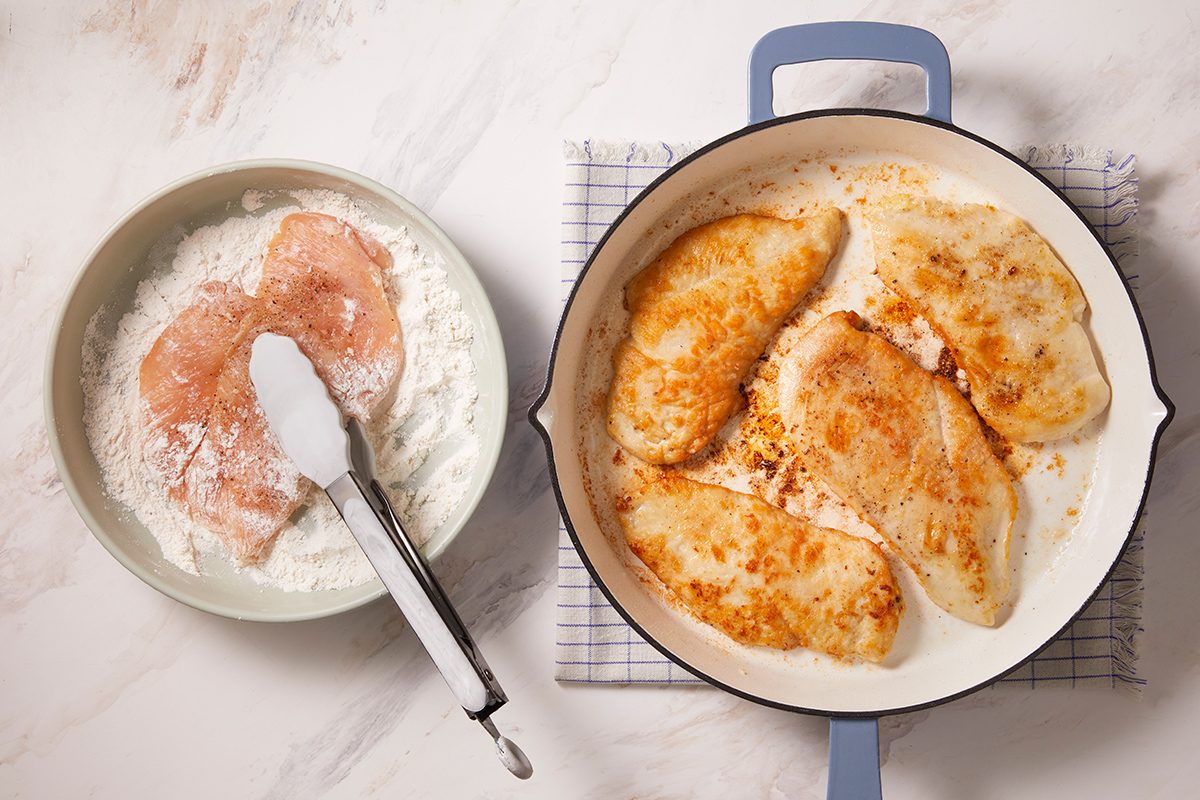 Chicken breasts are coated in flour in a bowl with tongs, while several coated chicken breasts cook and brown in a white skillet on a stovetop, set on a checkered towel.