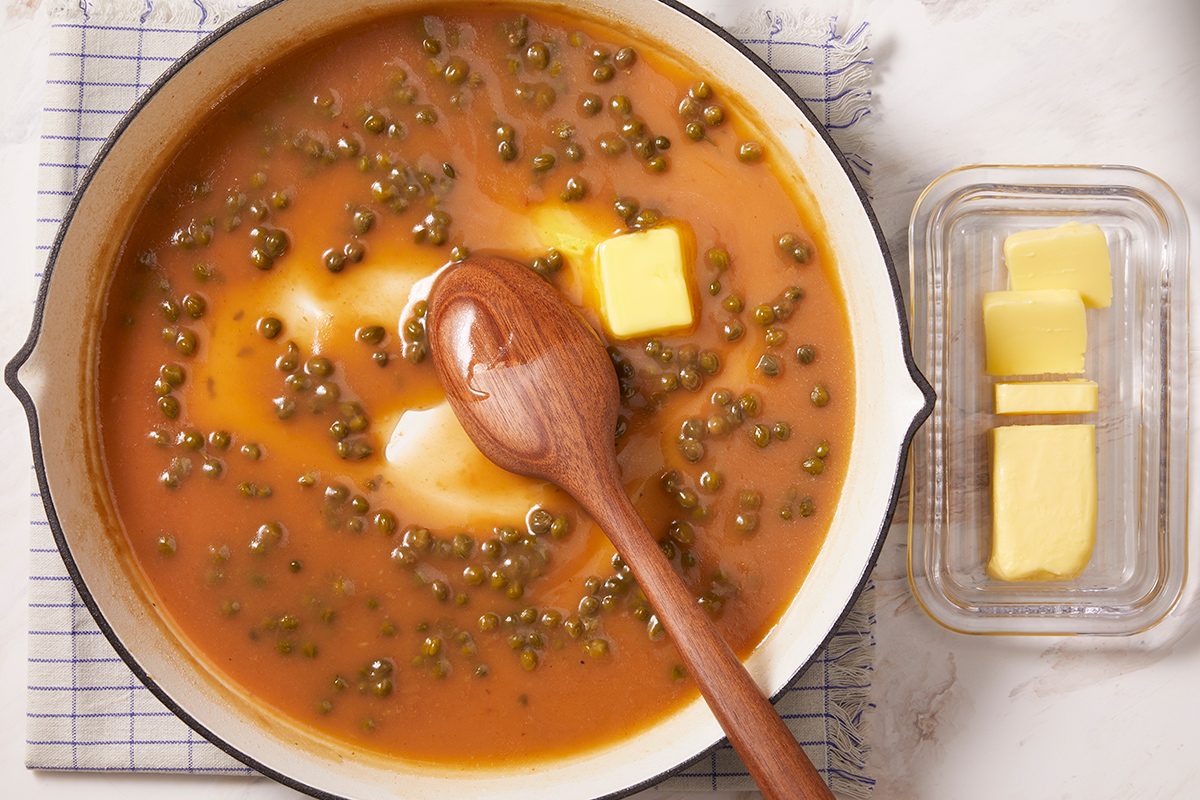 A pot of lentil soup with a red-orange broth, green lentils, and several chunks of butter, stirred with a wooden spoon. A butter dish with slices of butter sits nearby on a white surface.