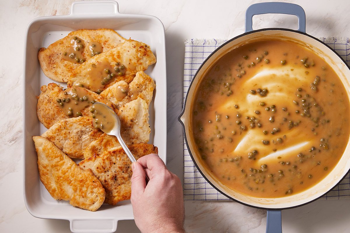 A hand spoons a caper-filled sauce over cooked chicken breasts in a baking dish, with more sauce in a saucepan nearby on a white marble surface.