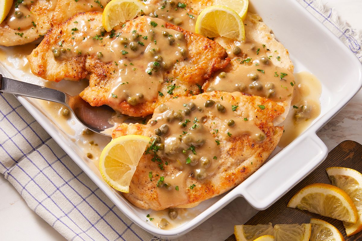 Golden chicken breasts topped with a creamy caper sauce, garnished with chopped herbs and lemon slices, served in a white baking dish with a serving spoon on the side.