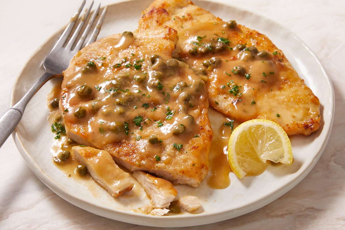 Two pieces of chicken piccata topped with caper and lemon sauce, garnished with chopped herbs, served on a white plate with a lemon wedge and a fork.