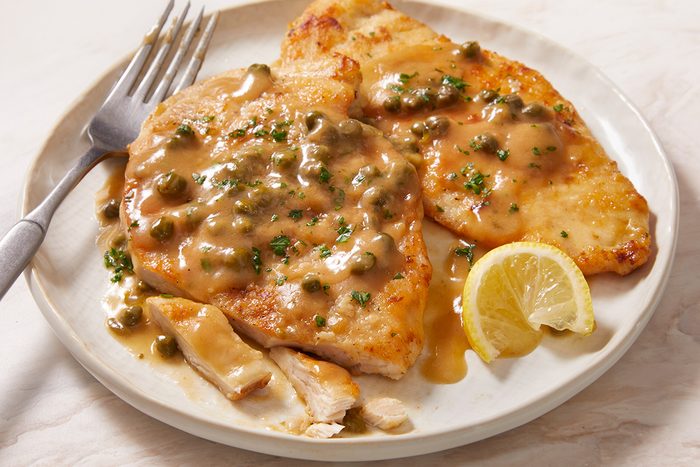 Two pieces of chicken piccata topped with caper and lemon sauce, garnished with chopped herbs, served on a white plate with a lemon wedge and a fork.