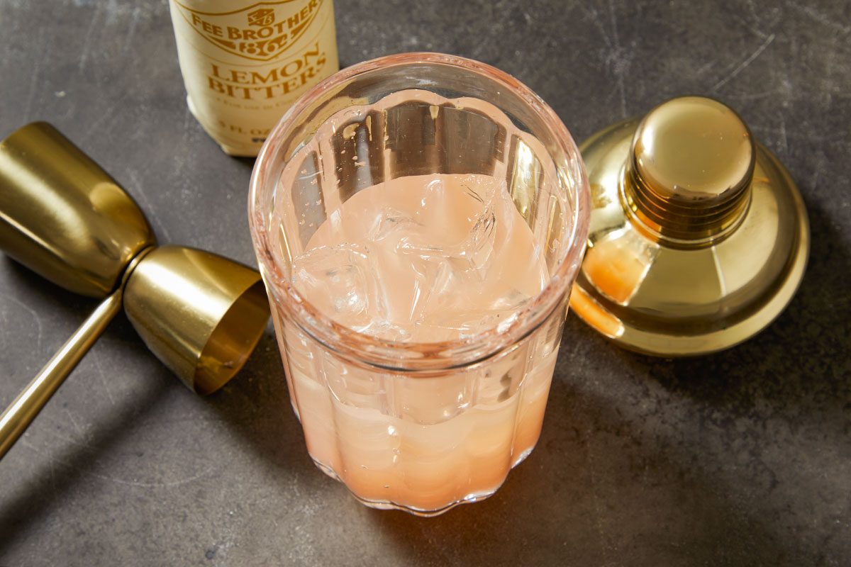 Overhead horizontal image of ice cubes in a mixing glass surrounded by bar tools and cocktail ingredients for a French Blonde Cocktail. A gold jigger and bottle of Lillet Blanc rest nearby on a dark surface, highlighting preparation for the cocktail. This step-by-step preparation image emphasizes the measured ingredients and method before mixing.