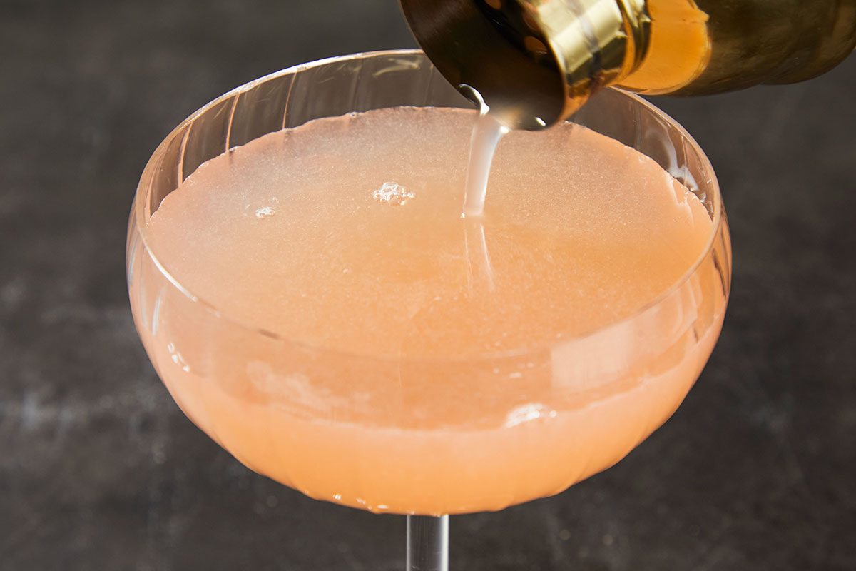 Horizontal overhead action image of French Blonde Cocktail being poured into a coupe glass. The pale pink cocktail streams from a bottle into the chilled glass, capturing the step-by-step method of assembling the drink. The soft citrus hue highlights the fresh grapefruit juice and Lillet Blanc.