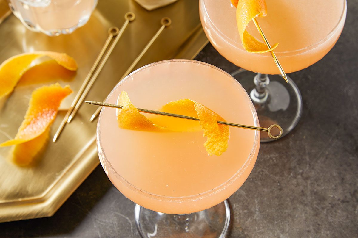 Horizontal push and pulls image featuring two French Blonde Cocktails in coupe glasses garnished with grapefruit peel twists. Styled with gold cocktail picks and citrus wedges on a dark surface, this image highlights entertaining and presentation of the finished drink.