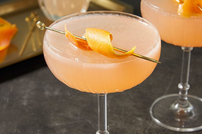 Horizontal push and pulls close-up image of two French Blonde Cocktails in coupe glasses with grapefruit peel garnish. The pale pink citrus cocktail is highlighted against a dark background, emphasizing texture, color, and refined presentation.