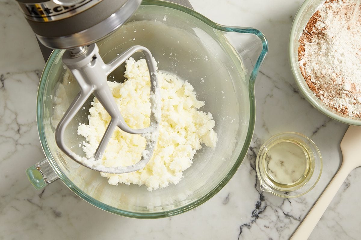 A stand mixer with a paddle attachment mixes creamed butter and sugar in a glass bowl. Nearby, a bowl of flour mixture, a small bowl of oil, and a spatula are on a marble countertop.