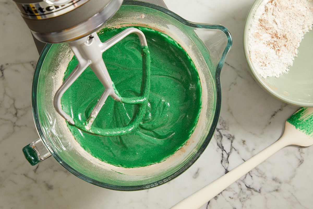 A mixing bowl with bright green batter and a stand mixer's paddle attachment, sitting on a marble countertop. Nearby, a plate with flour and a spatula with green batter are visible.