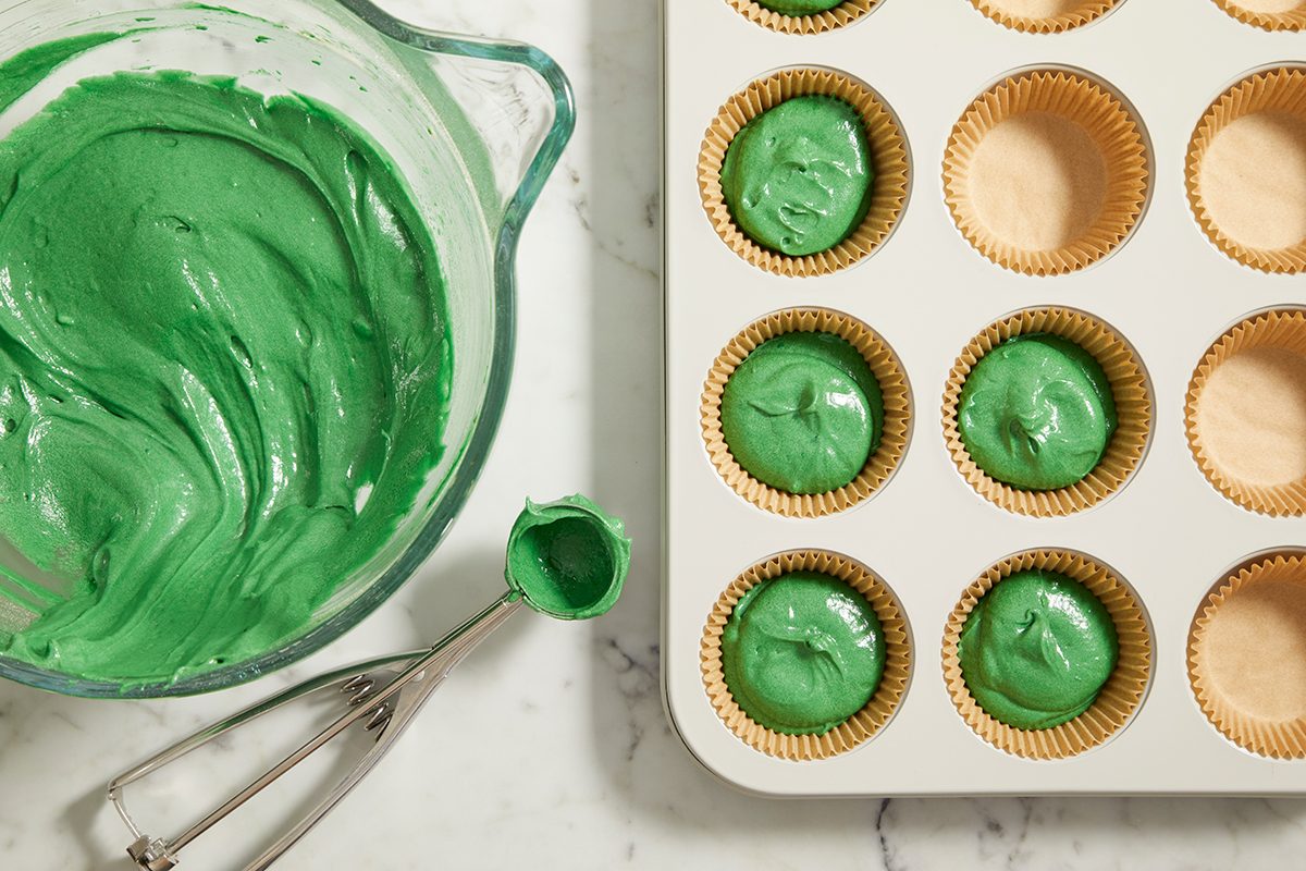 A mixing bowl with green cupcake batter sits next to a muffin tin lined with paper cups, some filled with green batter and some empty, on a marble countertop.