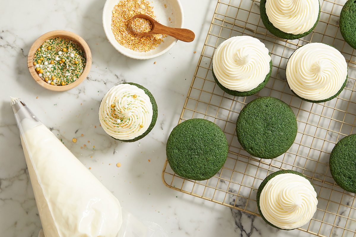Cupcakes with green tops, some frosted with white swirls, are on a cooling rack and marble countertop. Nearby are a piping bag with frosting and small bowls of sprinkles and sugar for decorating.