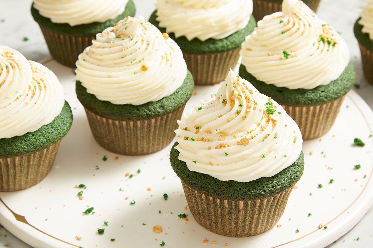 A group of green cupcakes with swirled white frosting, topped with sprinkles, arranged on a white plate.