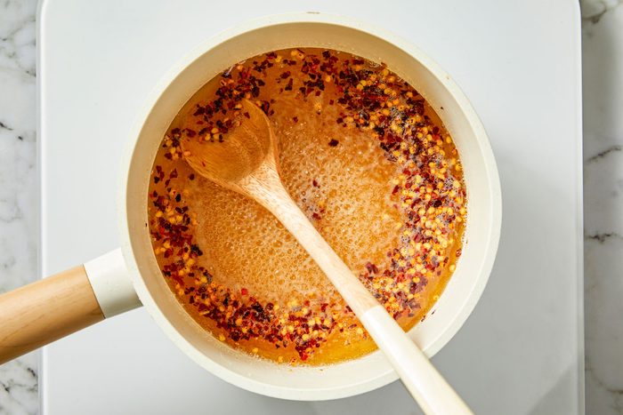 Overhead shot of a white saucepan on a stovetop; filled with simmering orange-brown liquid with chili flakes; stirred with a wooden spoon;