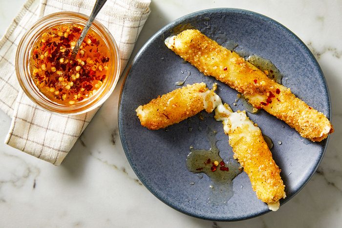 Overhead shot of Hot Honey Fried Mozzarella; three golden, crispy mozzarella sticks drizzled with chili-flecked hot honey on a dark grey plate; melted cheese oozes from the centers; a jar of spicy honey with a spoon sits on a folded kitchen towel beside the plate;