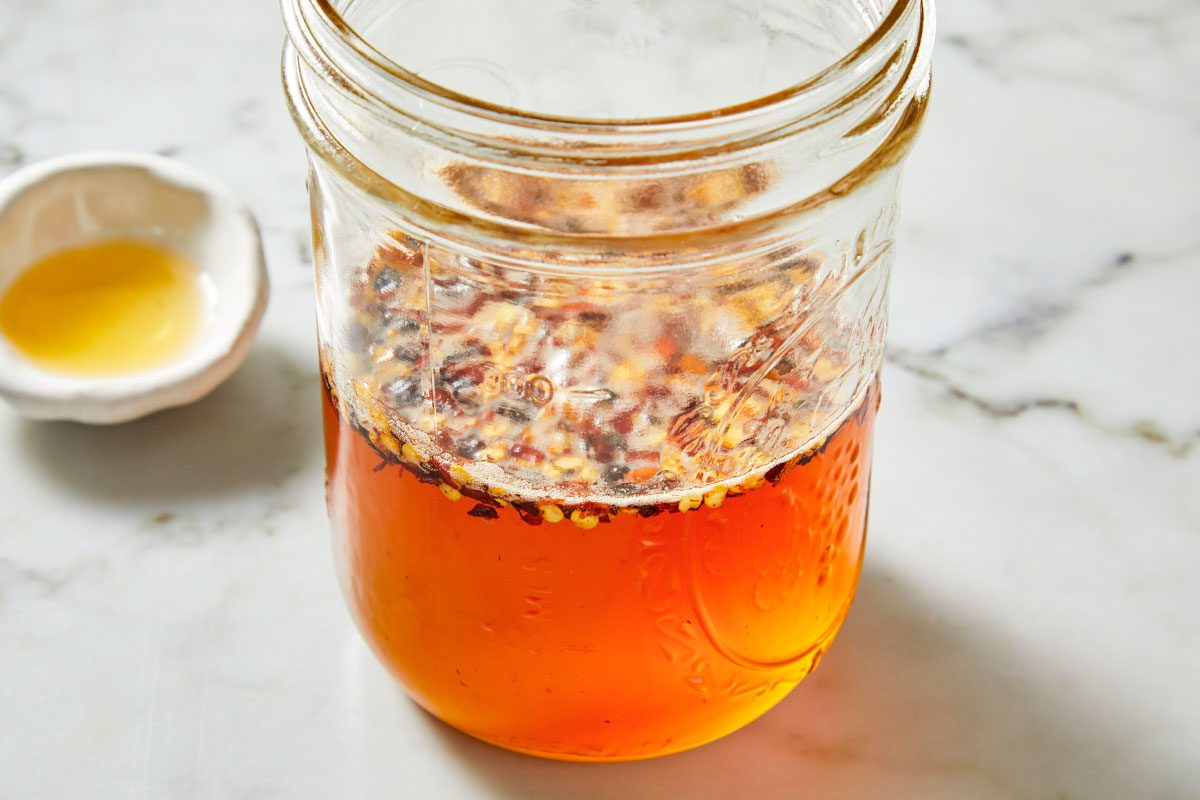Closeup shot of a glass jar filled with homemade chili oil; vibrant red chili flakes suspended in bright orange oil; set on a white marble surface with a small dish of yellow oil blurred in the background;