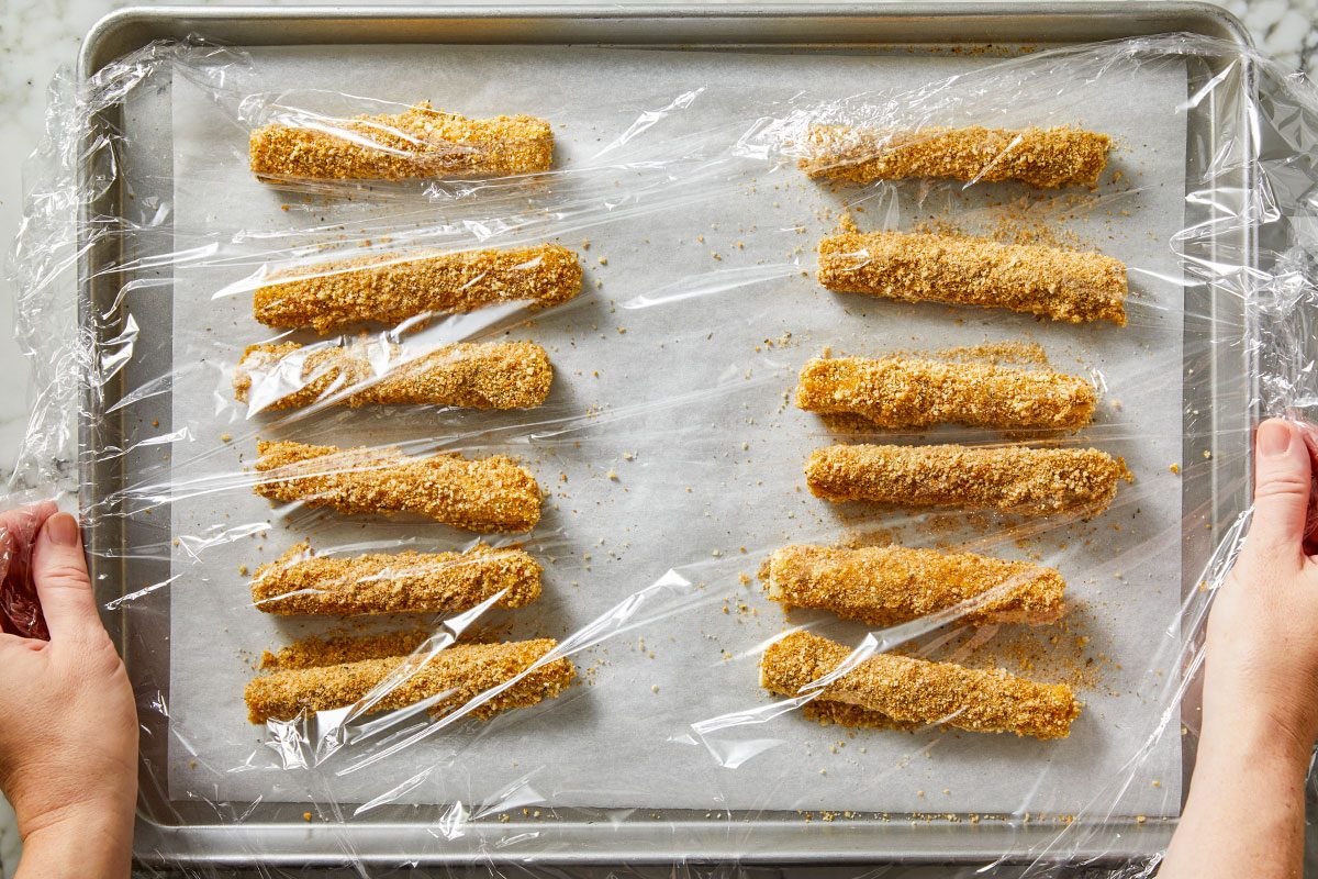 Overhead shot of breaded cheese sticks arranged in neat rows on a parchment-lined baking sheet; hands pull plastic wrap over the tray to cover the coated sticks;