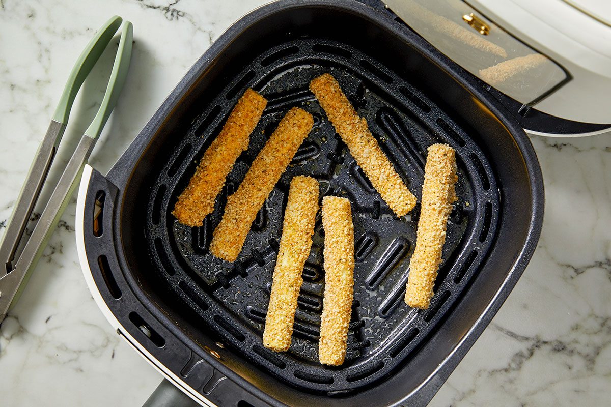 Overhead shot of breaded cheese sticks arranged in a single layer inside an air fryer basket; the sticks are evenly spaced on the dark nonstick surface; tongs rest on the marble countertop beside the fryer;