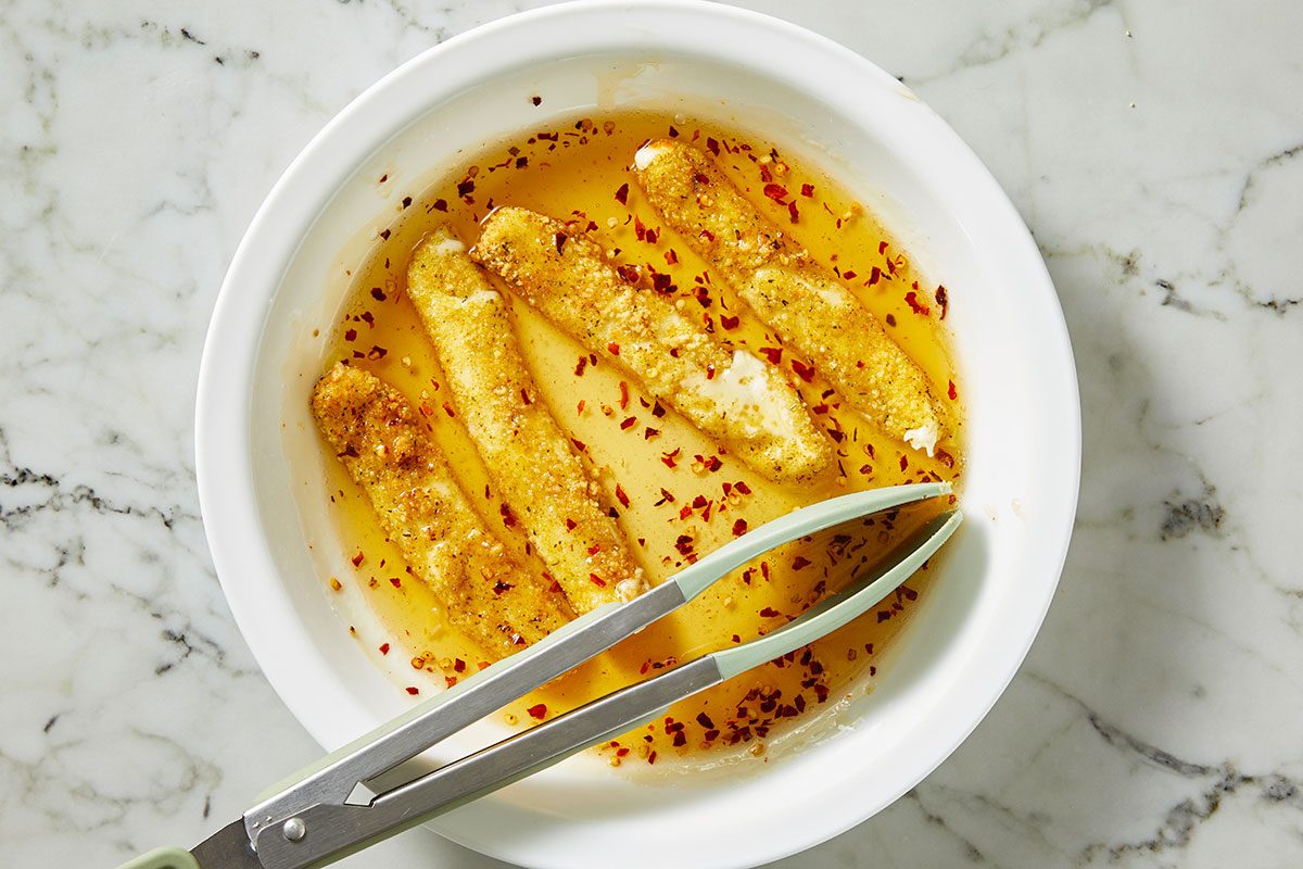 Overhead shot of air-fried cheese sticks in a white bowl; coated in chili oil with visible red pepper flakes; a pair of tongs rests in the bowl as the sticks are turned and coate;