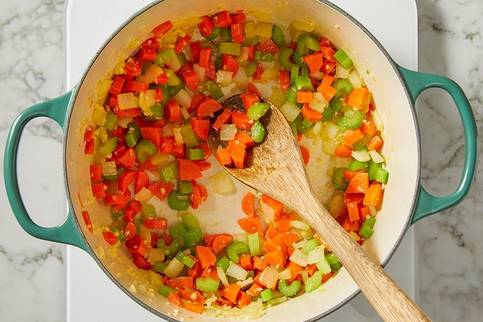 Overhead, horizontal step-by-step image of chopped onion, carrot, and celery sautéing in olive oil in a light-colored Dutch oven, with a wooden spoon stirring the vegetables. This image captures the early cooking stage of Italian Penicillin Soup, showcasing aromatic vegetables softening in oil for a homemade chicken soup base.