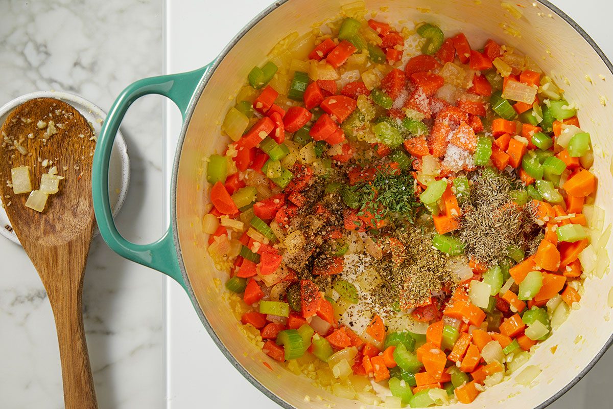 Overhead view of sautéed onion, carrot, and celery being seasoned with dried herbs, salt, and pepper in a Dutch oven. This step-by-step image highlights seasoning the vegetable base for Italian Penicillin Soup before adding broth.