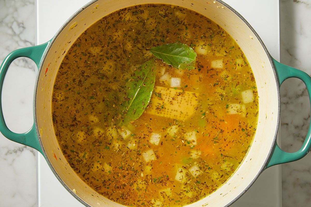 Overhead, horizontal image of chicken broth added to seasoned vegetables in a Dutch oven, with bay leaf and Parmesan rind visible. This step shows the soup transitioning from sautéed vegetables to simmering broth for Italian Penicillin Soup.