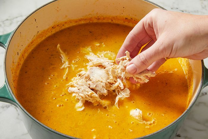 Overhead shot of shredded cooked chicken breast being added to a creamy soup base in a Dutch oven. This step highlights building protein and heartiness in Italian Penicillin Soup.