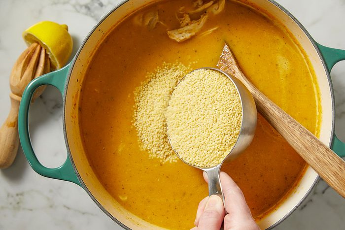 Overhead view of uncooked pastina being poured into simmering soup in a Dutch oven. This image captures the step of adding pasta to Italian Penicillin Soup for texture and heartiness.