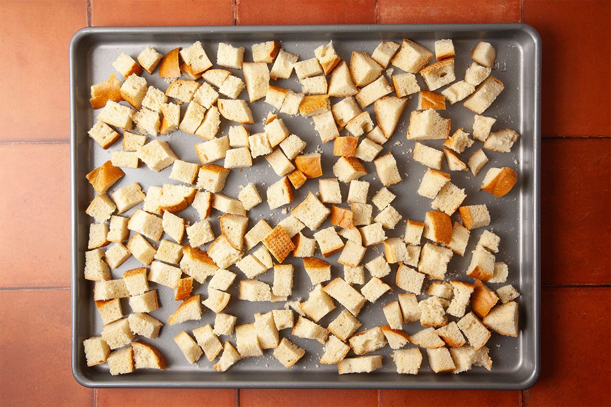Overhead step-by-step image of cubed Italian bread arranged on a sheet pan before baking. The evenly cut 1-inch cubes are spread across a metal baking sheet on a warm-toned tile surface, preparing for homemade croutons to top Marry Me Chicken Soup.