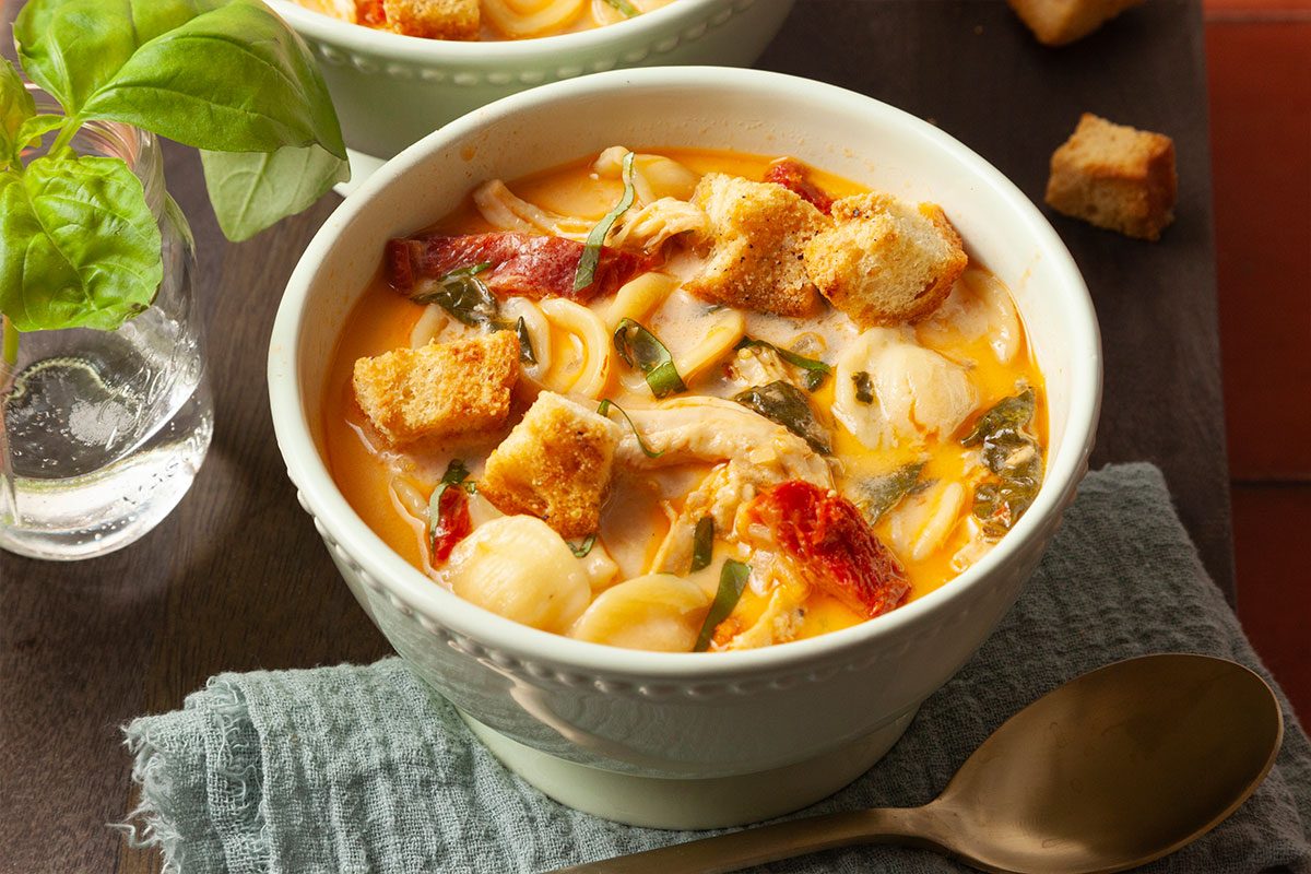 Push-and-pulls styled close-up overhead image of Marry Me Chicken Soup highlighting tender chicken, orecchiette pasta, wilted spinach, sun-dried tomatoes, and crispy croutons in a creamy tomato broth.