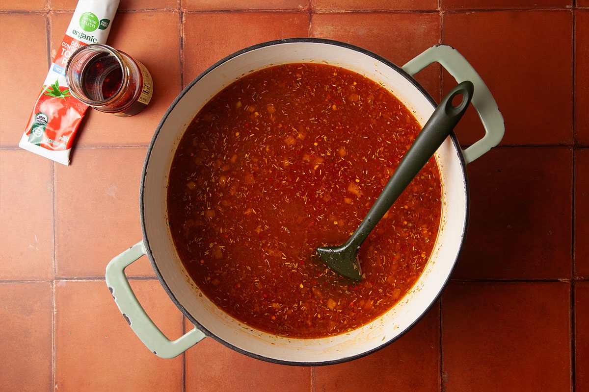 Overhead image of chicken broth simmering with tomato paste and julienned sun-dried tomatoes in a Dutch oven. The soup base deepens in color as seasonings are incorporated.