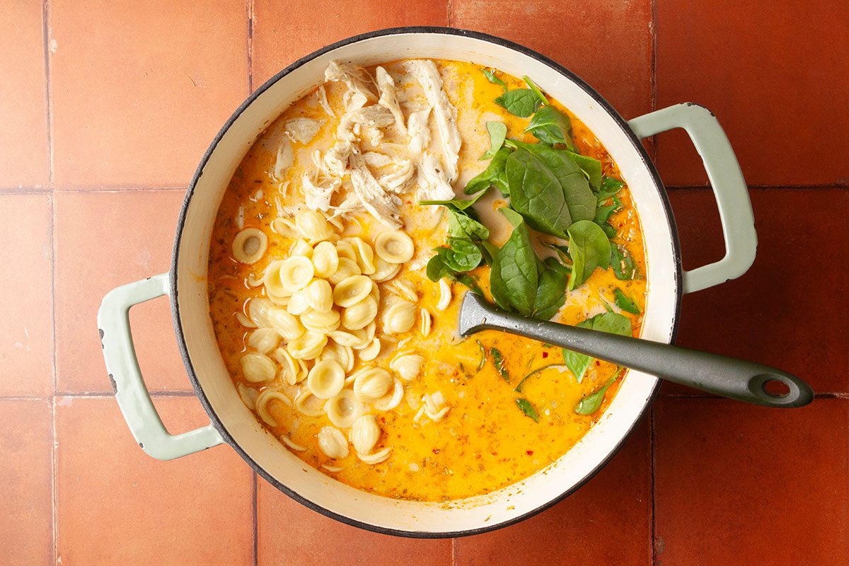 Overhead step-by-step image of orecchiette pasta, shredded chicken, and fresh baby spinach added to the creamy tomato broth. The soup begins to take on its hearty final form.