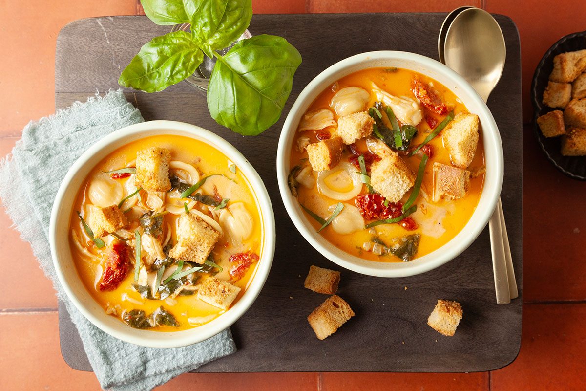 Beauty shot styled overhead image of two bowls of Marry Me Chicken Soup garnished with basil leaves and croutons. The creamy tomato broth, orecchiette, chicken, and spinach are visible in a cozy serving presentation.