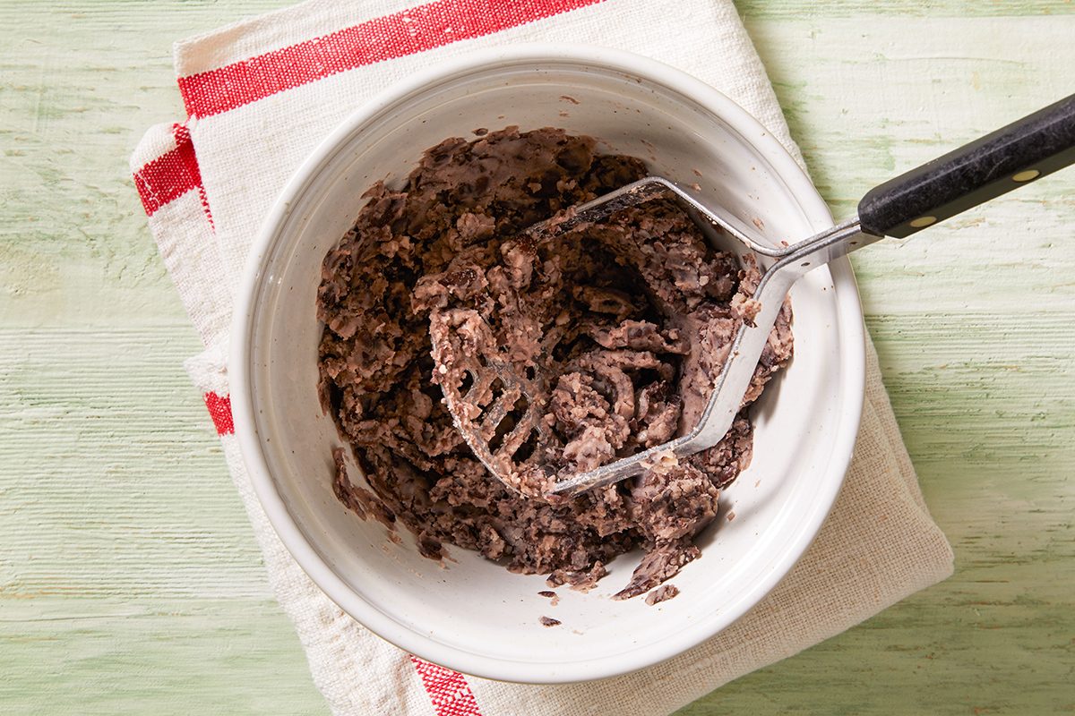 A white bowl containing mashed black beans sits on a folded white kitchen towel with red stripes; a metal potato masher rests inside the bowl.