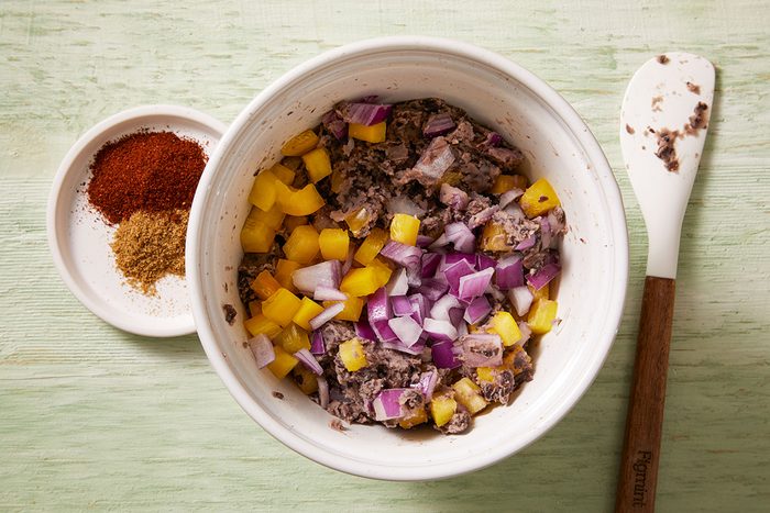 A bowl containing chopped yellow bell pepper, red onion, and black beans sits next to a white spoon. A small plate with two piles of ground spices is placed beside the bowl on a light green surface.
