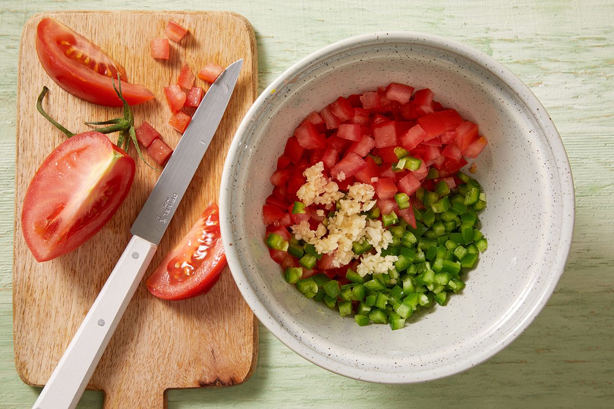 A cutting board with sliced tomatoes and a knife sits beside a bowl containing diced tomatoes, diced green bell peppers, and minced garlic on a light green surface.