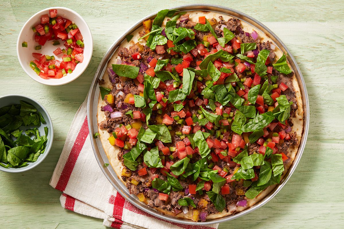 A pizza topped with ground meat, chopped tomatoes, spinach, and red onions on a metal tray. Nearby are small bowls of salsa and fresh spinach on a white and red dish towel over a light green surface.