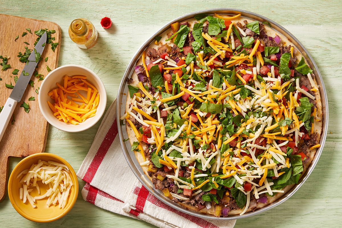 A pizza topped with shredded cheese, spinach, onions, and peppers on a baking tray, next to bowls of extra cheese and chopped herbs on a wooden board with a knife, all set on a light-colored surface.