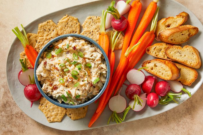Million-Dollar Dip served in a bowl surrounded by crackers, baguette slices, carrots and radishes.