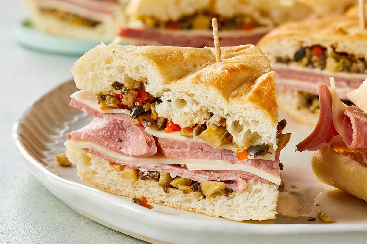 Close-up shot of a mini muffuletta sandwich with layers of meat, cheese, and olive salad on a white plate;
