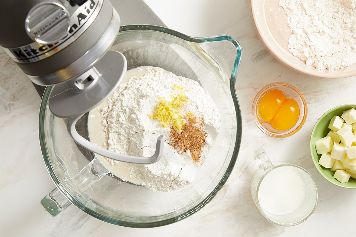 Overhead view of measured ingredients for New Orleans King Cake arranged around a stand mixer bowl. Yeast, warm liquids, sugar, butter, egg yolks, lemon zest, and spices are prepared for mixing, illustrating the beginning stages of dough preparation.