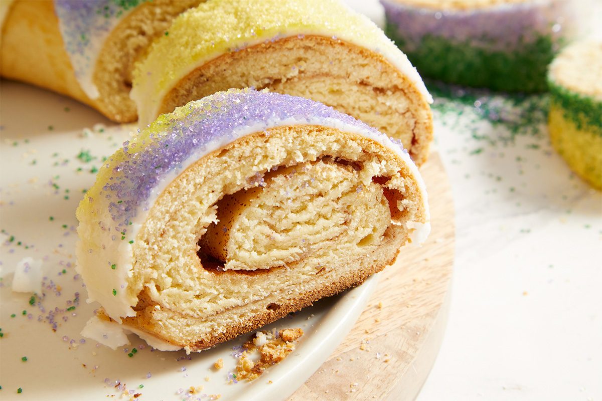 Tight overhead horizontal close-up of a sliced piece of King Cake, showcasing the spiral cinnamon filling and soft interior beneath the colorful sugar topping.