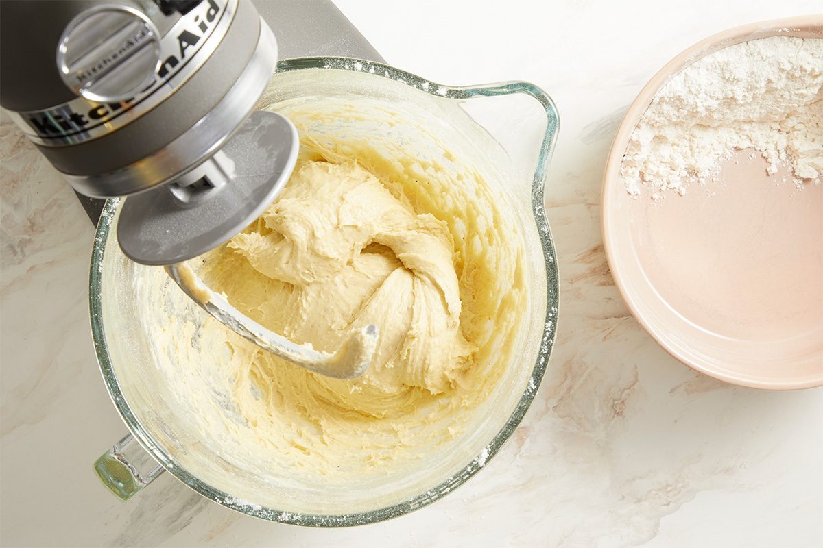 Overhead image of King Cake dough being mixed in a stand mixer bowl. The enriched dough appears smooth and creamy as ingredients are incorporated, highlighting the early mixing stage of this traditional New Orleans dessert.