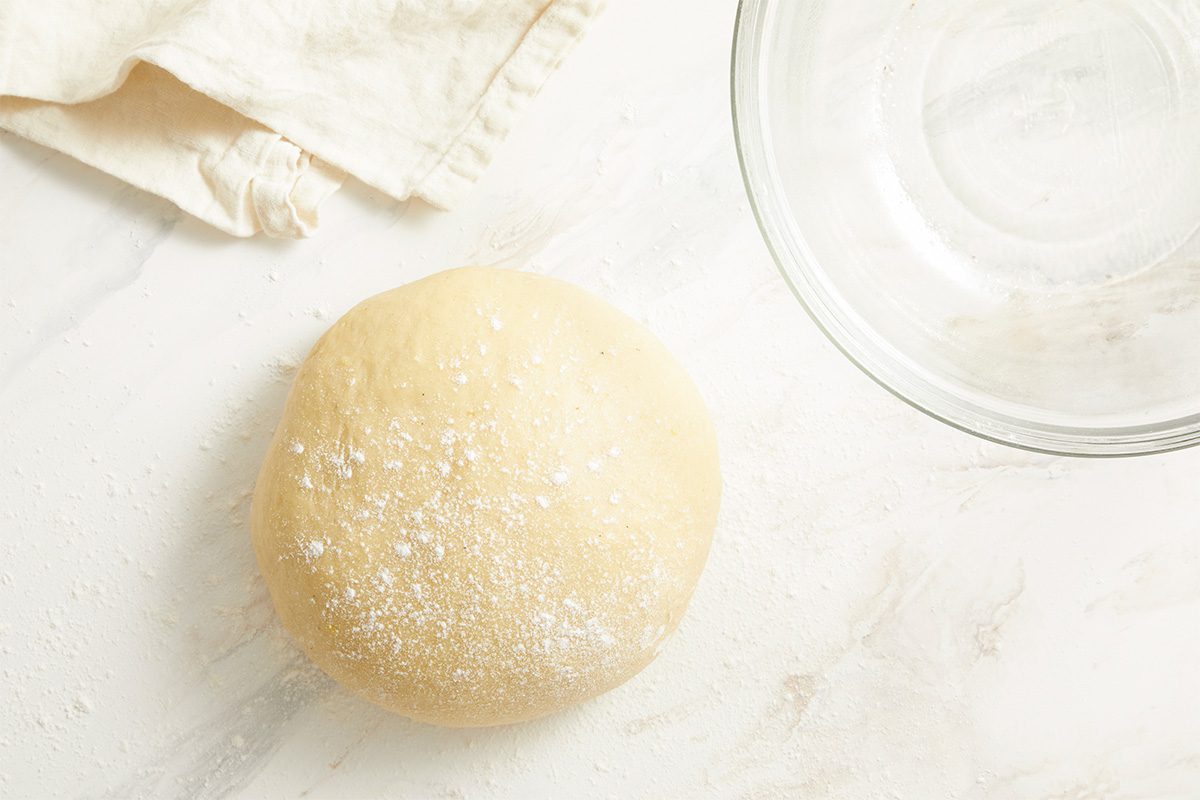 Overhead view of a smooth round ball of King Cake dough resting on a lightly floured surface, indicating the completion of kneading and readiness for shaping or rising.