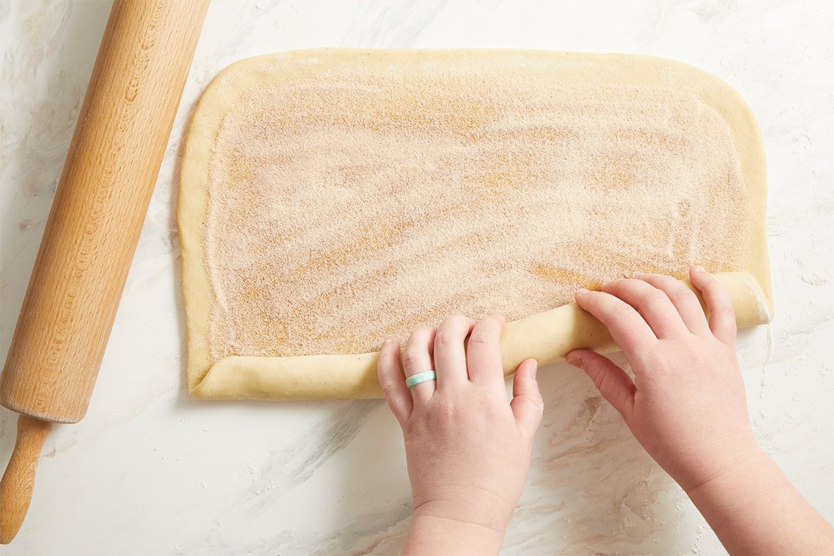 Overhead action shot of hands rolling King Cake dough into a rectangle on a lightly floured surface with a rolling pin, preparing the dough for filling and shaping.