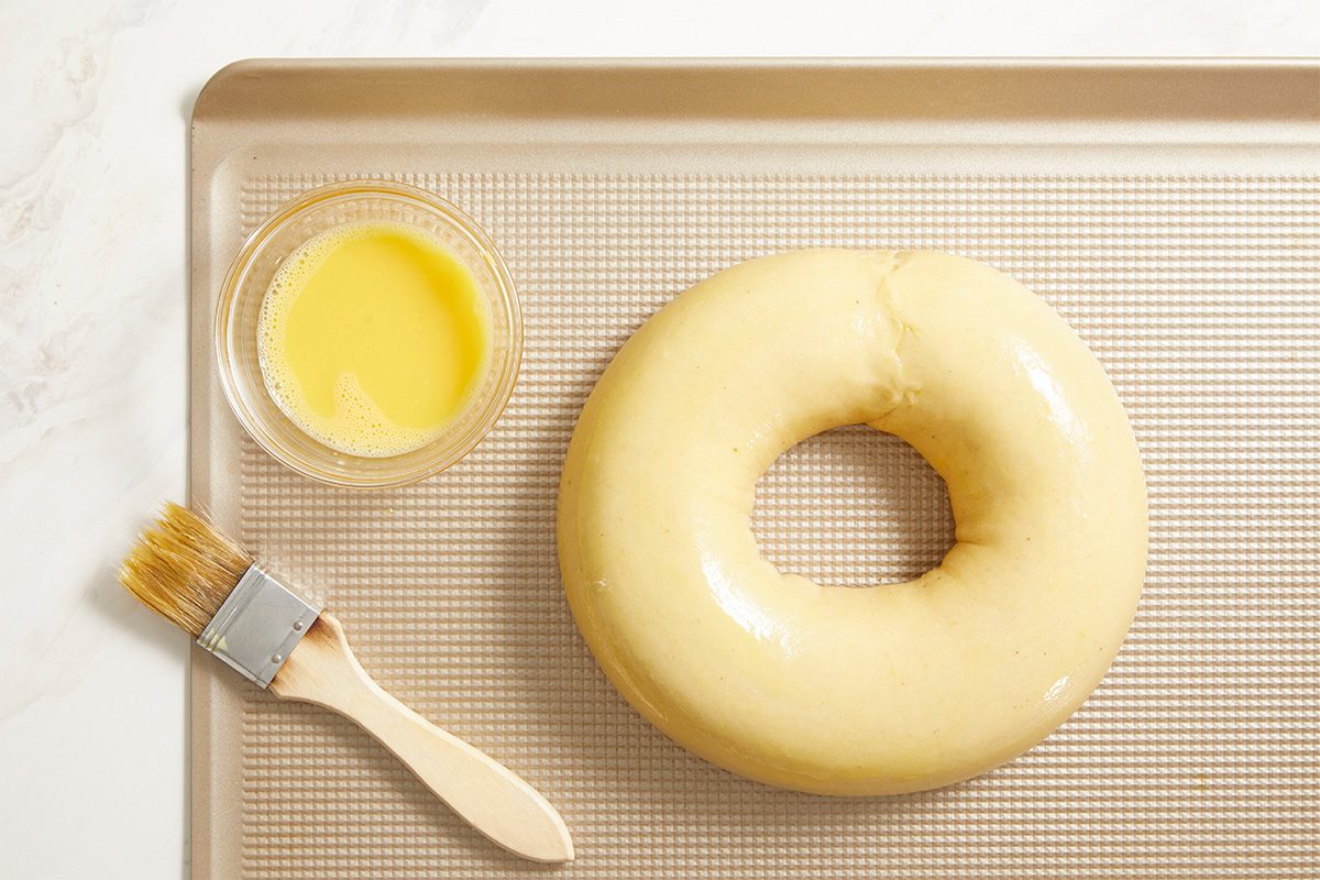 Overhead image of shaped King Cake dough formed into a ring on a baking sheet. A pastry brush and beaten egg sit nearby, showing the dough prepared for egg wash before baking.