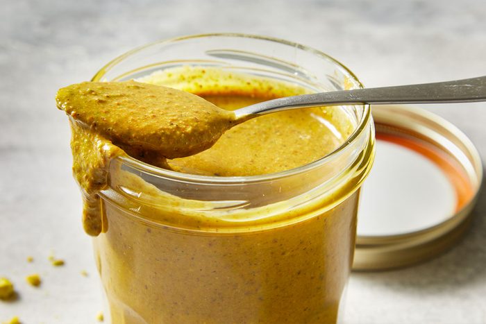 Horizontal close-up of a spoon lifting creamy Pistachio Butter from a glass jar, highlighting its thick, velvety texture and spreadable consistency.